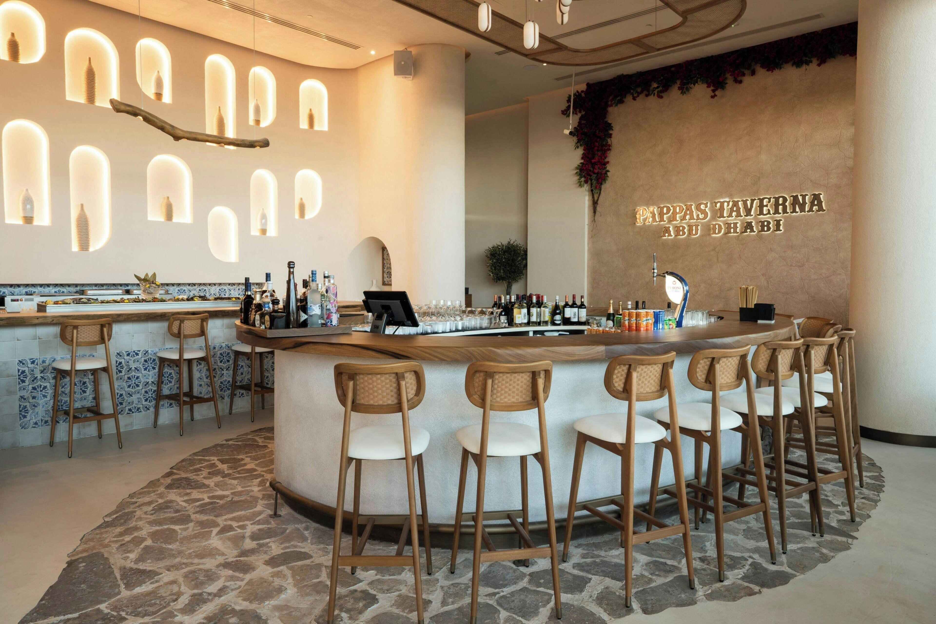 A bar area with a central counter, multiple stools, and a wall-mounted sign reading "Cappas Taverna Abu Dhabi".