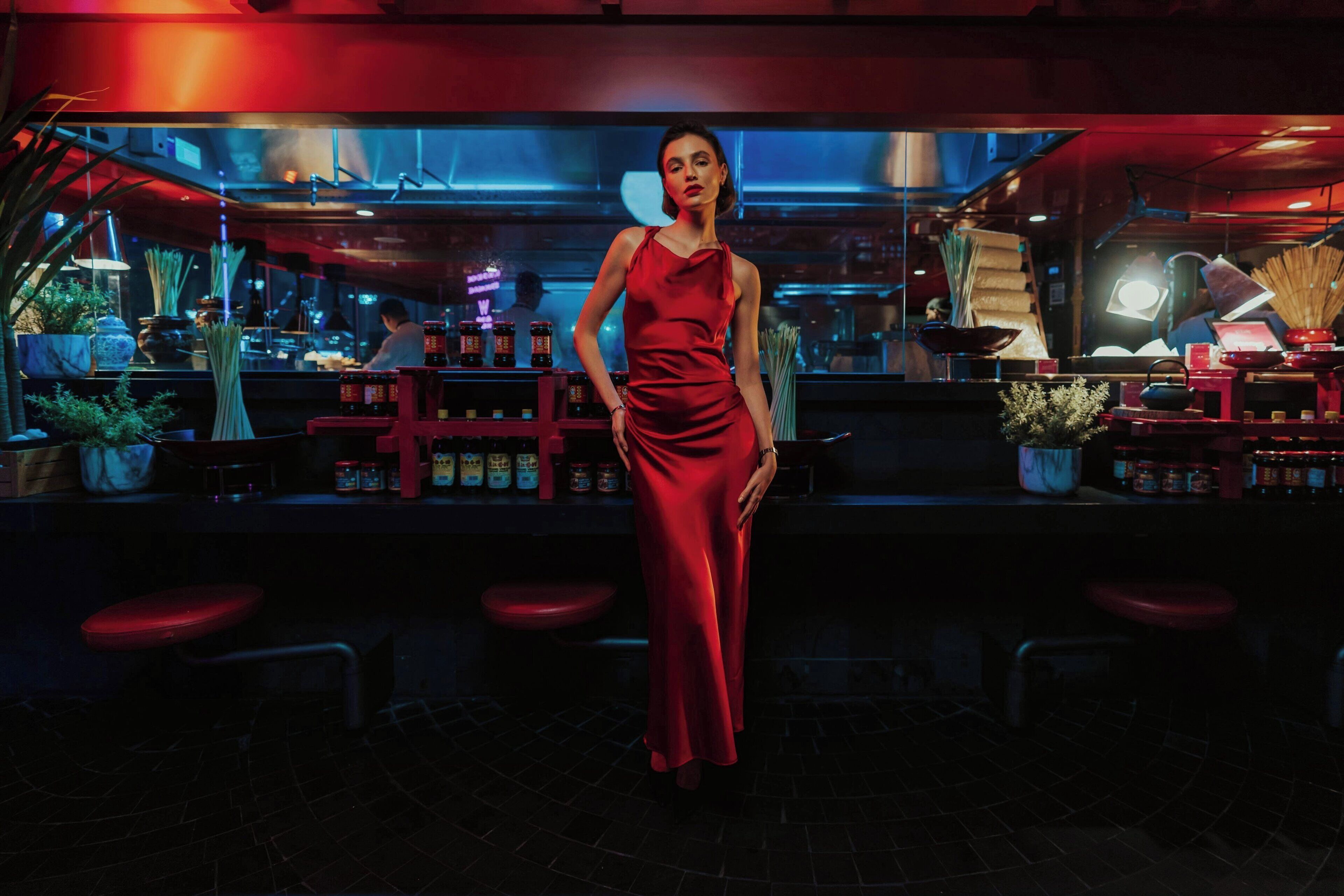 Uma mulher de vestido vermelho está em pé num bar com pouca luz, com banquetas e prateleiras cheias de garrafas.