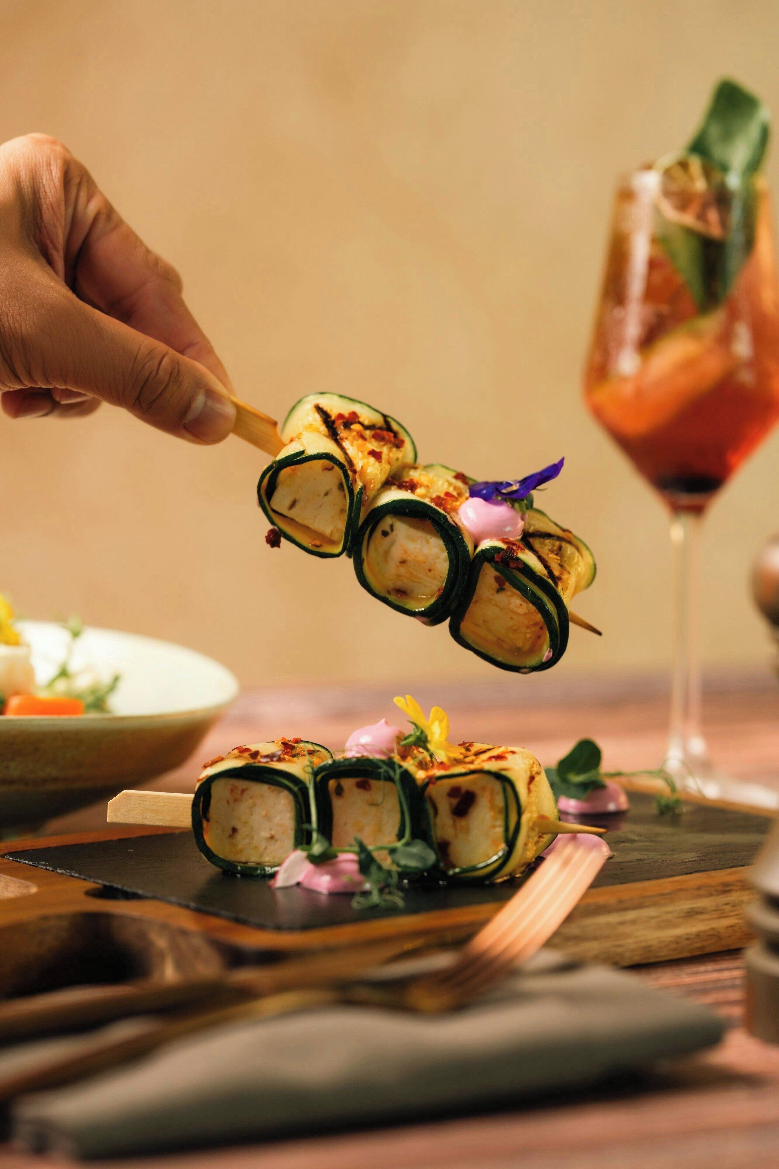 A hand holding a sushi roll with a decorative garnish, placed on a wooden board with a glass of drink in the background.