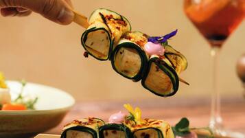 A hand holding a sushi roll with a decorative garnish, placed on a wooden board with a glass of drink in the background.