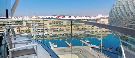 A modern cruise ship deck with a glass railing, a pool area, and a view of a marina with boats and a distinctive spherical building in the background.