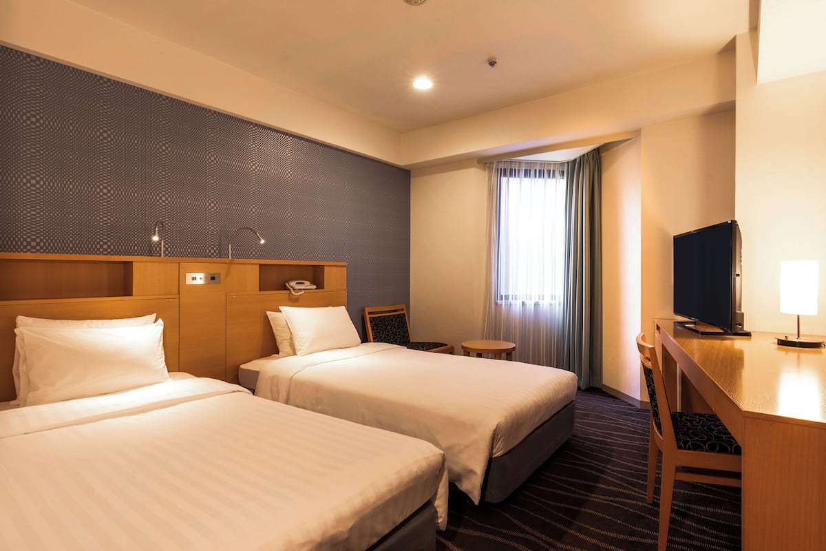 A hotel room with two beds, a desk with a chair, a television, and a window with curtains.