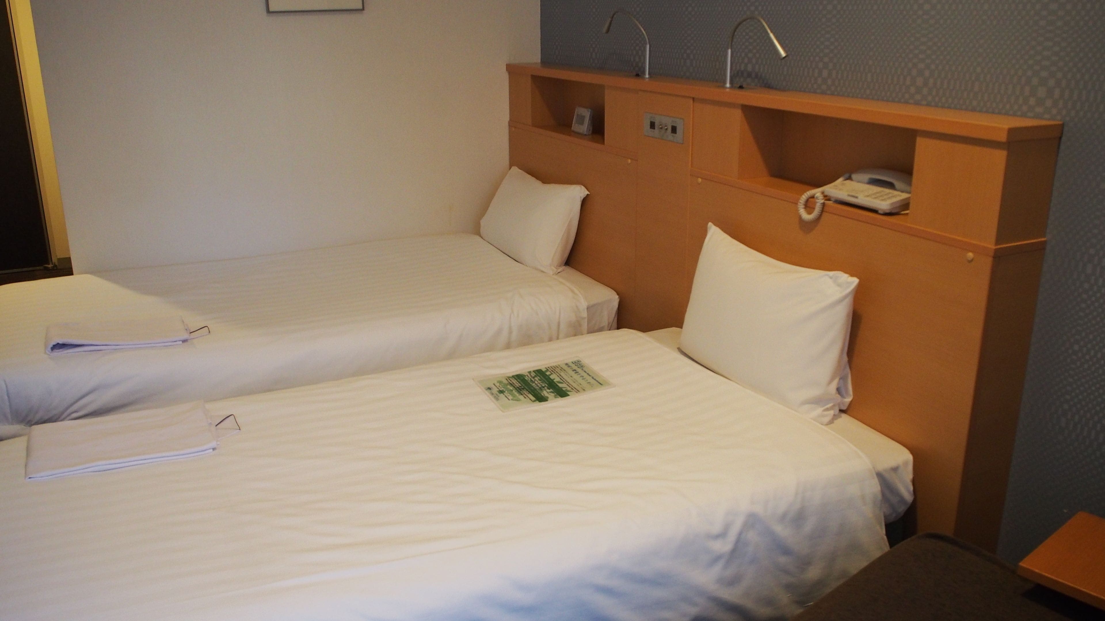 Two single beds with white linens, a wooden headboard with a telephone and a small shelf, and a wall-mounted light fixture.