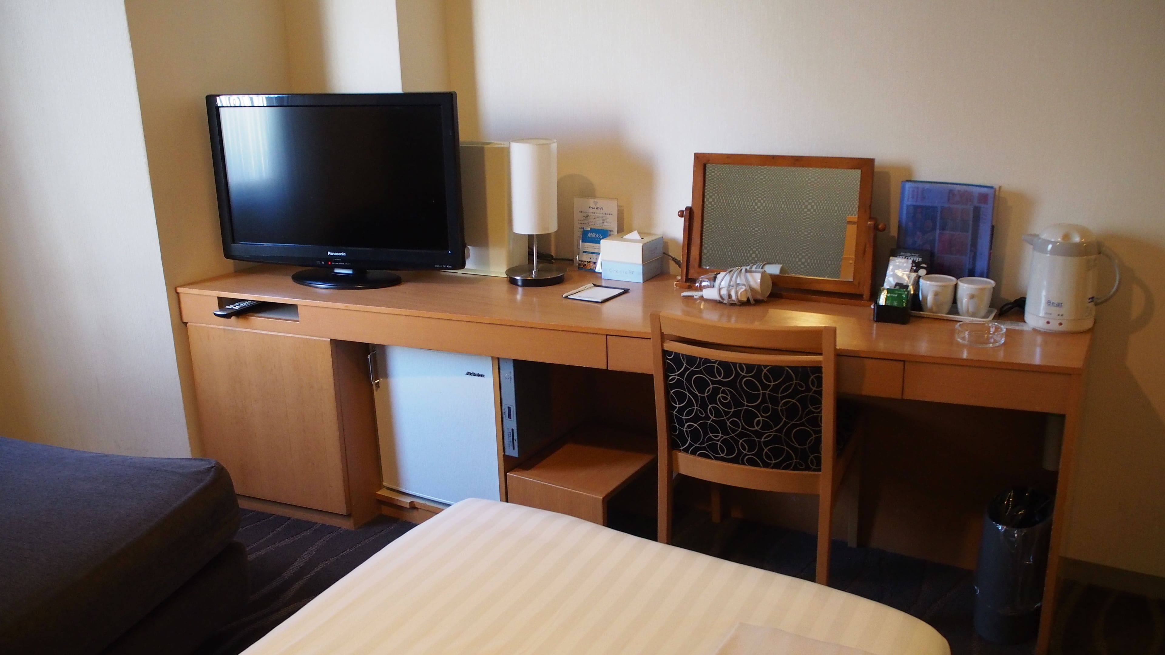 Una habitación de hotel con cama, escritorio con televisor, lámpara, refrigerador pequeño, silla y cafetera.