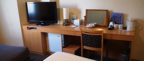 A hotel room with a bed, a desk with a television, a lamp, a small refrigerator, a chair, and a coffee maker.