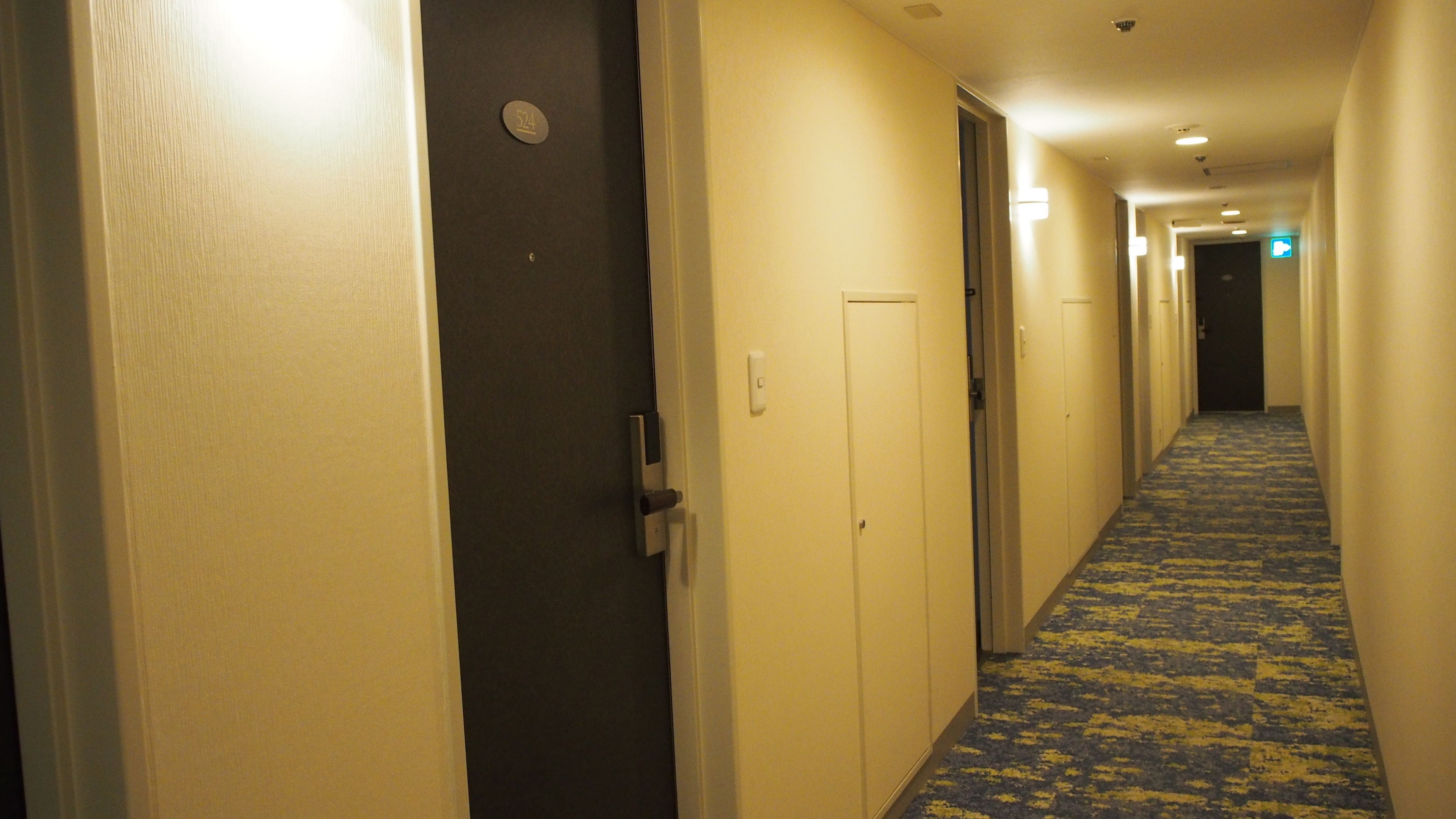 A hotel hallway with carpeted floors, doors, and a light switch on the wall.