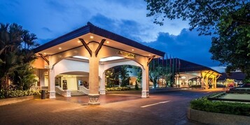 Entrada de hotel con un paseo techado, palmeras y una fachada bien iluminada.