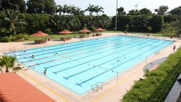 A large outdoor swimming pool with multiple lanes, surrounded by palm trees and shaded by red umbrellas.