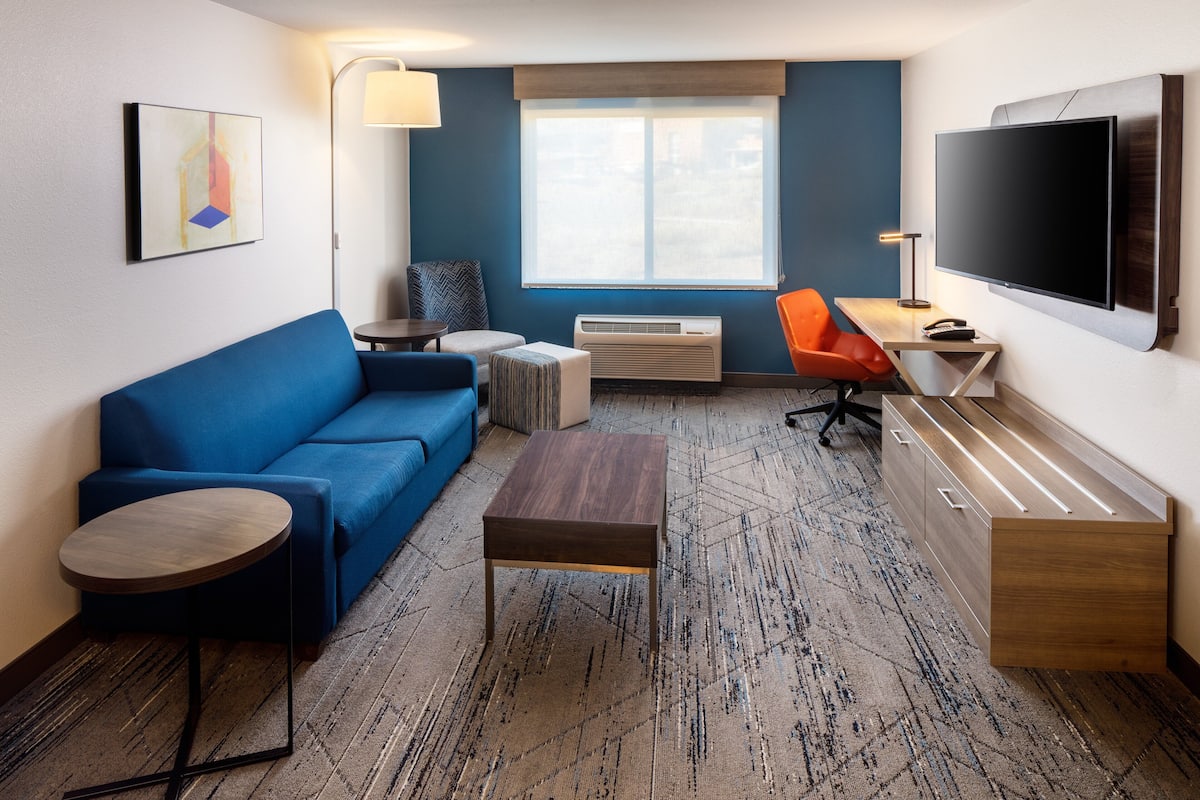 A modern hotel room with a blue sofa, a small round table, a wooden desk, a flat-screen TV, and a large window with a view.