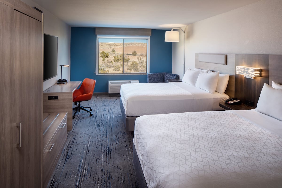 A hotel room with two beds, a desk, a chair, a large window with a desert view, and a wall-mounted lamp.