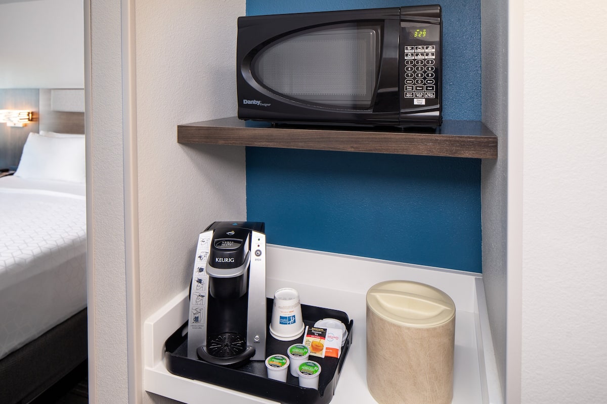 A hotel room with a microwave on a shelf, a coffee maker, and a tray with cups and condiments.