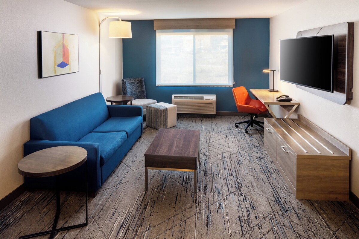 Une chambre d’hôtel moderne avec un canapé bleu, une petite table ronde, un bureau en bois, un téléviseur à écran plat et une grande fenêtre donnant sur un paysage agréable.