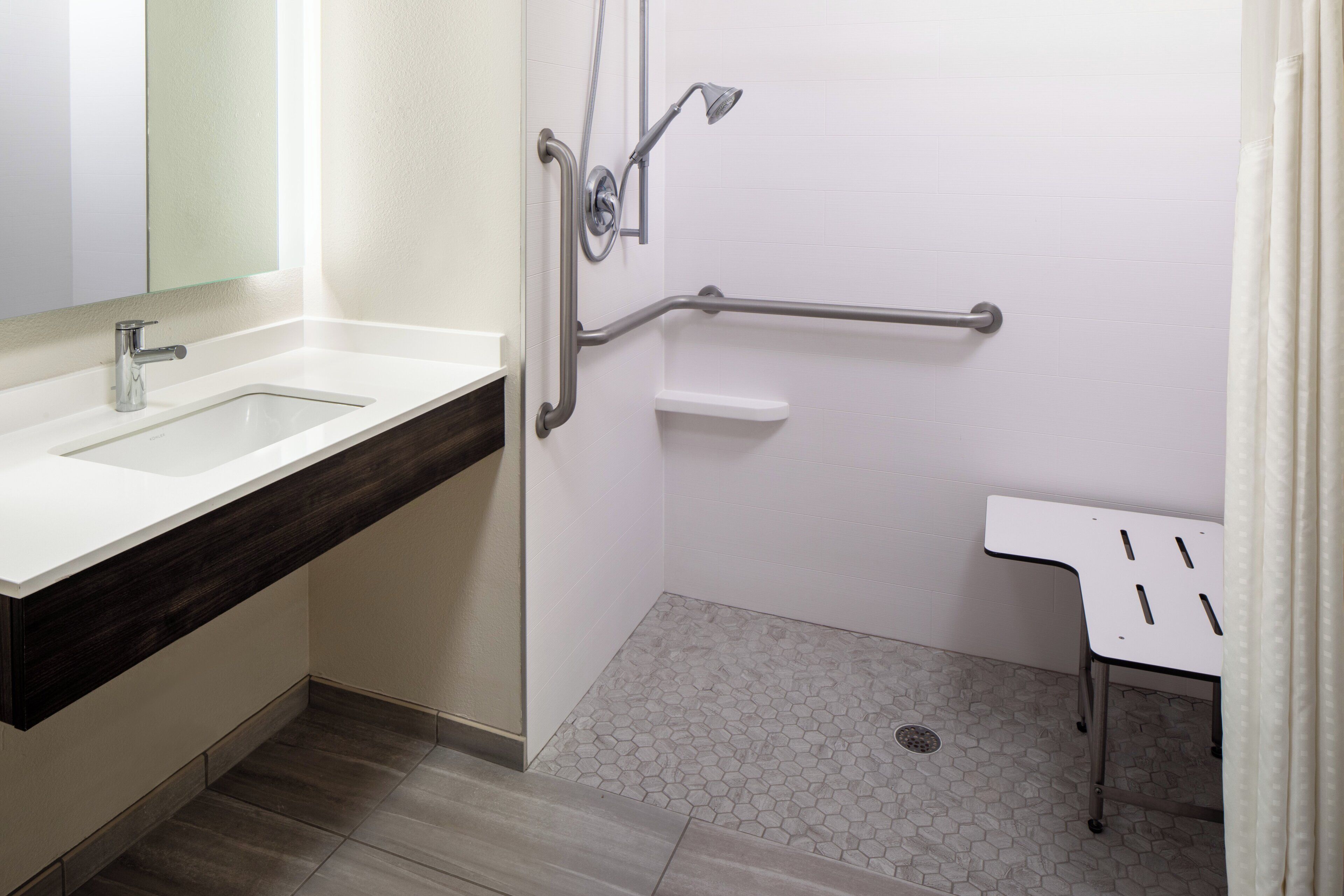 Un baño moderno con un lavamanos blanco, una ducha con barra de apoyo y una pequeña mesa blanca.