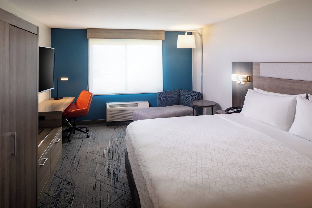 A hotel room with a large bed, a desk with a chair, a TV, a small table, and a window with blinds.