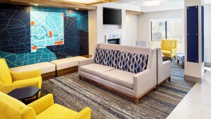 Un hall d’hôtel moderne avec un canapé, des fauteuils jaunes, un mur à motifs, un téléviseur à écran plat et une cheminée.
