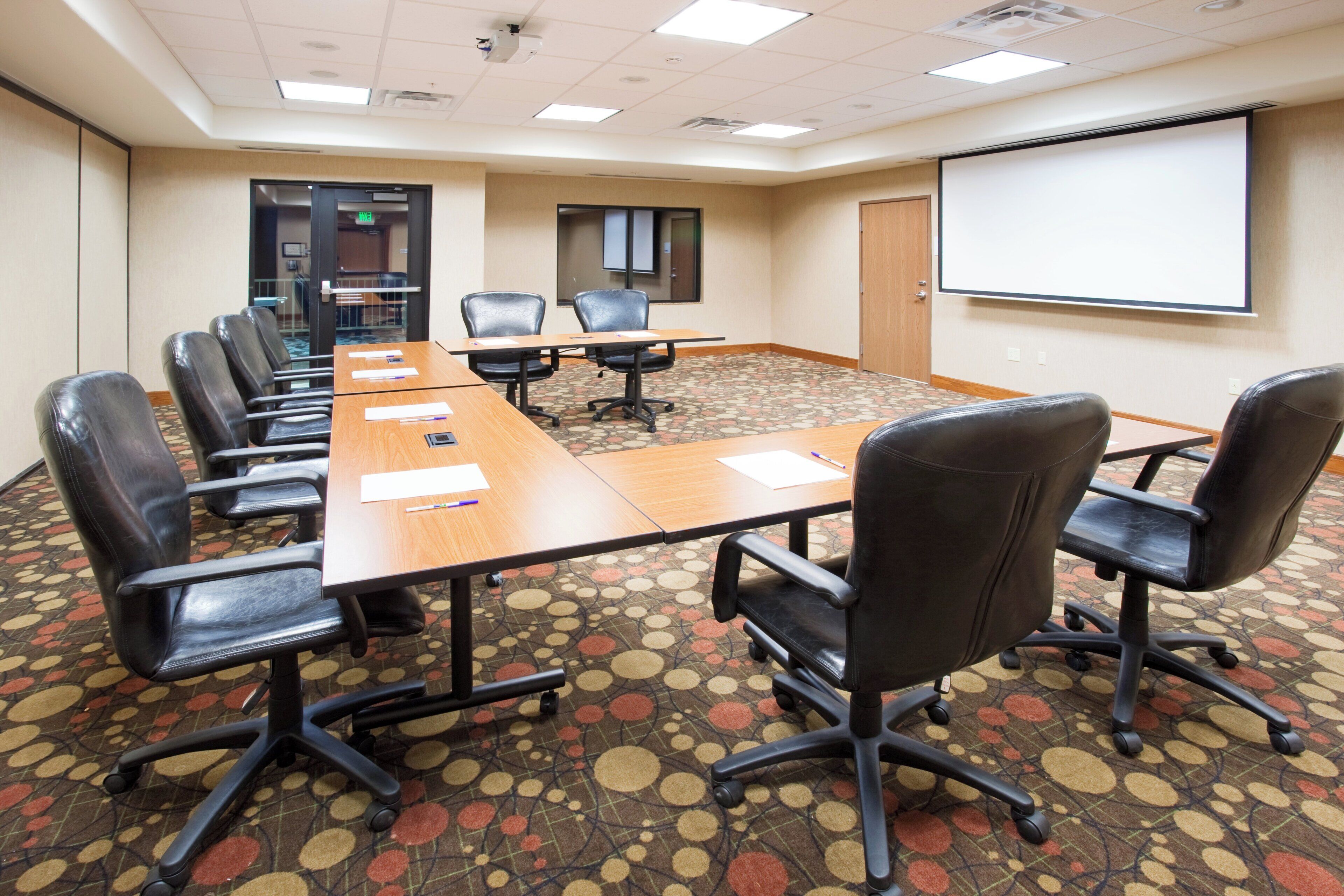 A conference room with a long table, multiple chairs, a projector screen, and a carpeted floor.