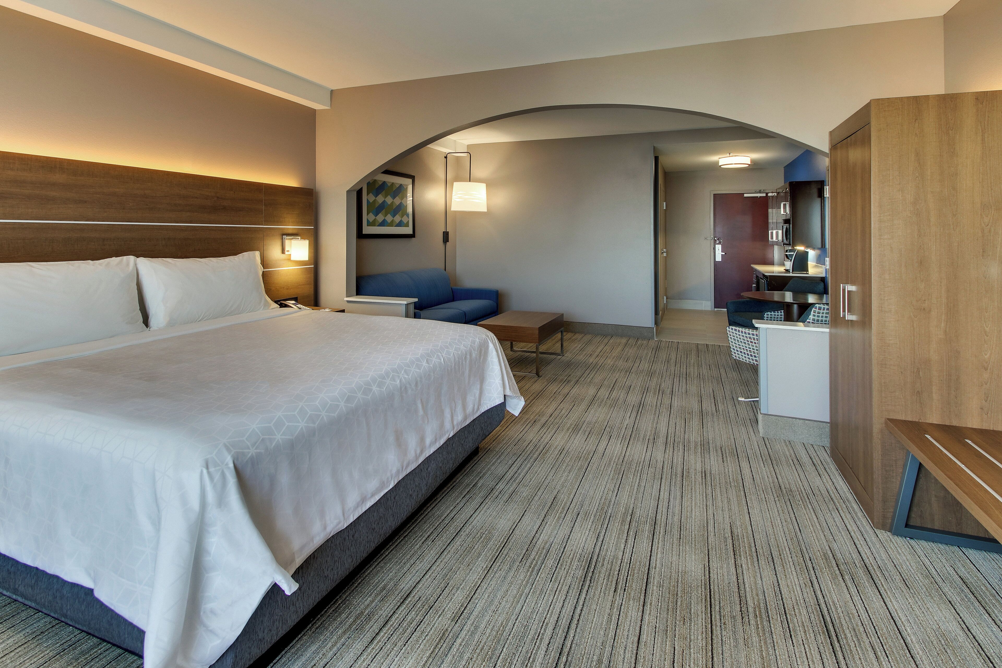 A hotel room with a large bed, a desk, a chair, a sofa, and a kitchenette area.