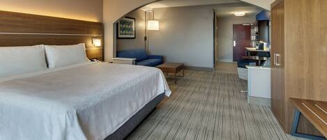 A hotel room with a large bed, a desk, a chair, a sofa, and a kitchenette area.