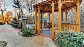 A wooden gazebo with a stone wall backdrop, a pool, and a paved walkway.