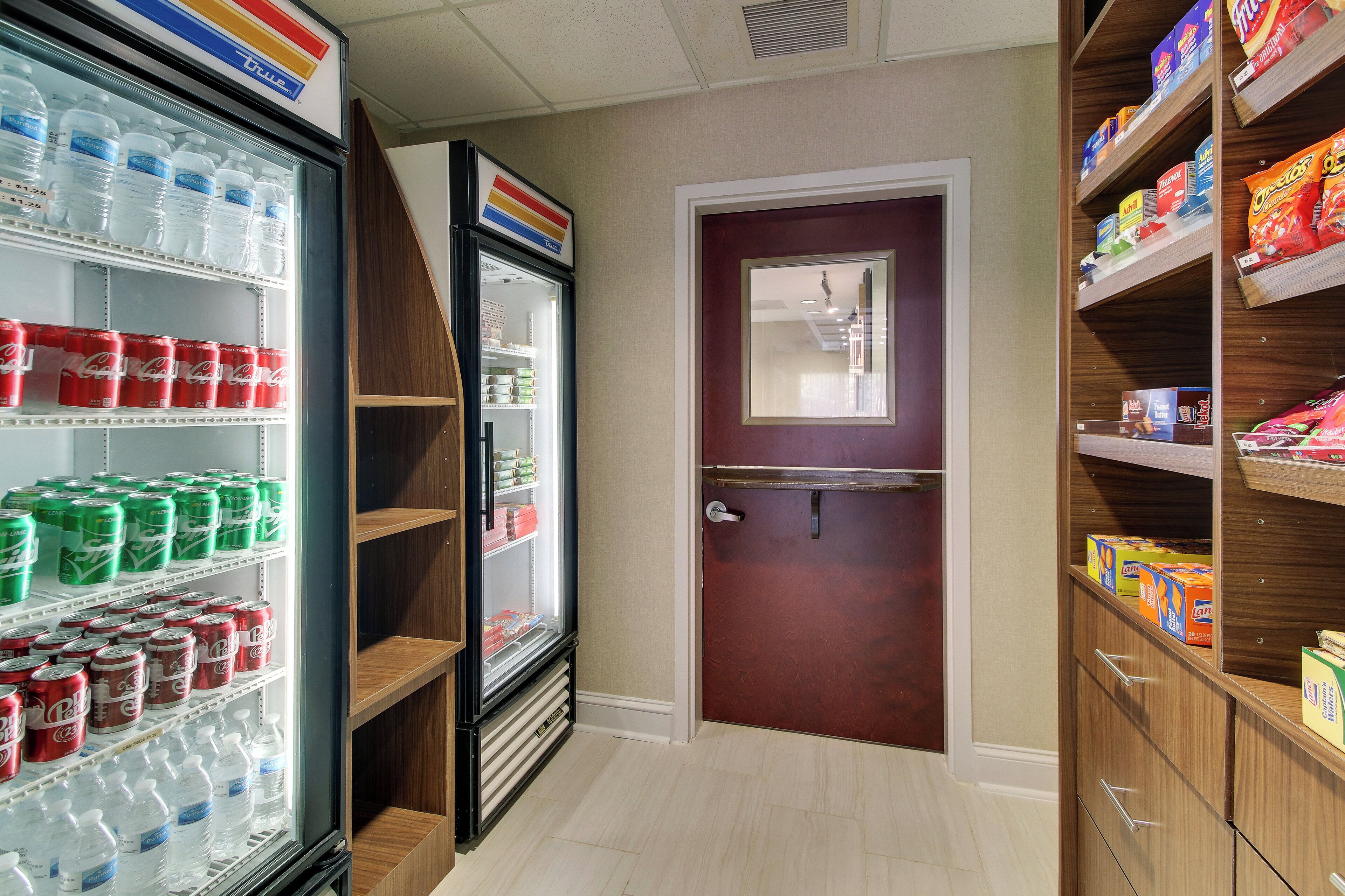 A hotel corridor with a refrigerator stocked with bottled water and soda, a wooden door, and shelves with snacks and other items.