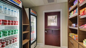 A hotel corridor with a refrigerator stocked with bottled water and soda, a wooden door, and shelves with snacks and other items.