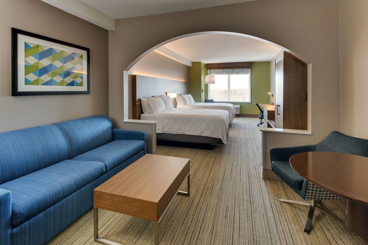 Une chambre d’hôtel avec un canapé bleu, une table basse en bois, un lit avec une literie blanche et un grand miroir qui reflète la pièce.
