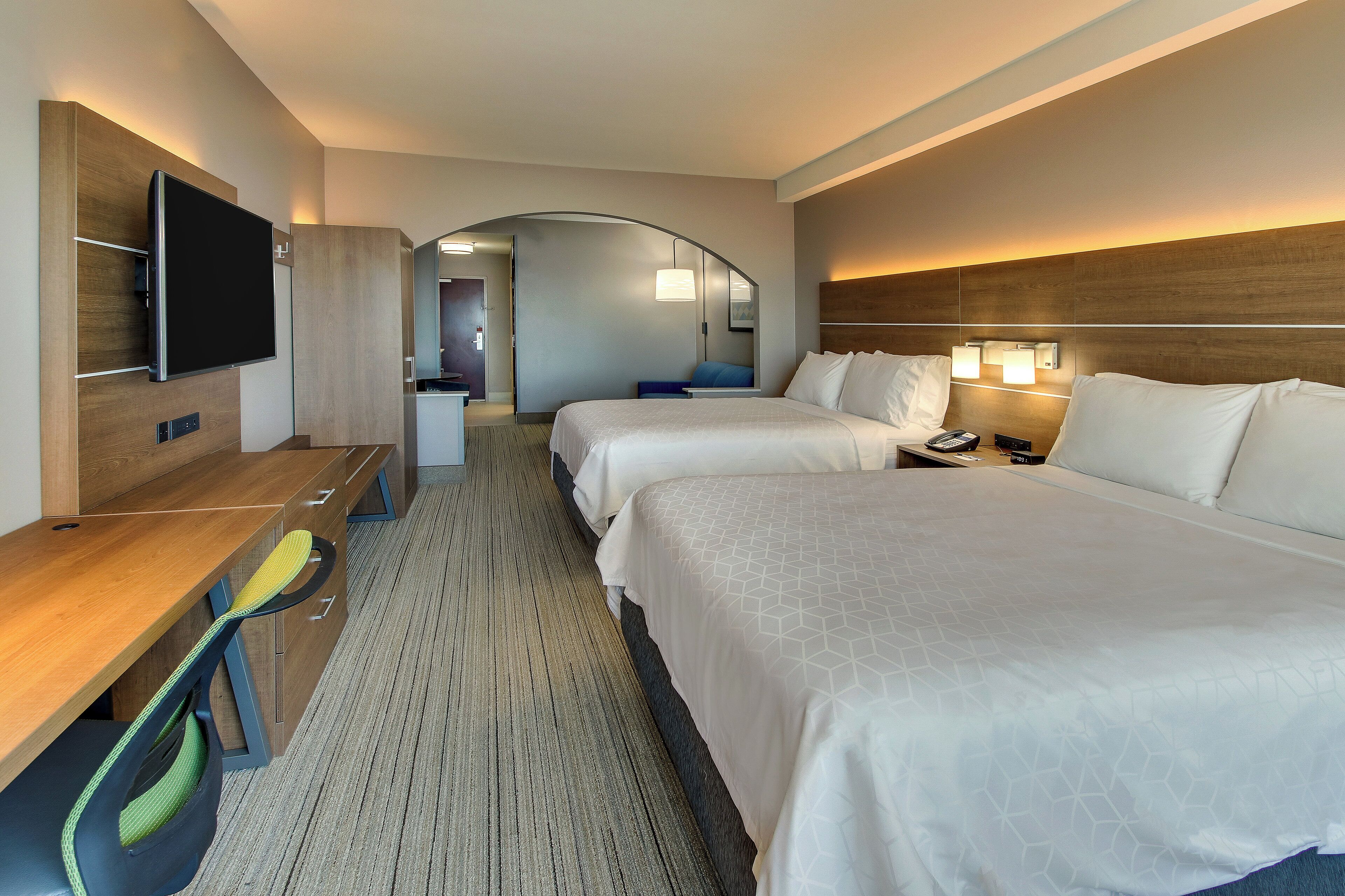 A hotel room with a desk, two beds, a TV, and a bathroom visible through an archway.