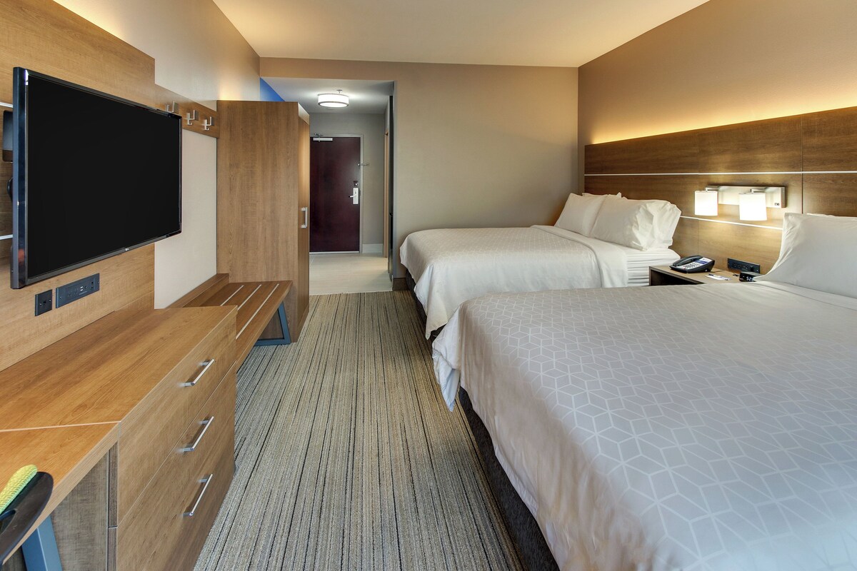 Habitación de hotel con televisión de pantalla plana, un escritorio de madera con cajones, dos camas con sábanas blancas, una mesita de noche con lámpara y suelo alfombrado.