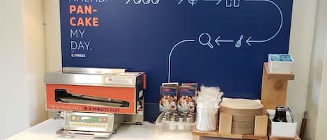 A kitchen area with a flat-screen TV displaying a breakfast menu, a pancake-making machine, and various breakfast items on the counter.