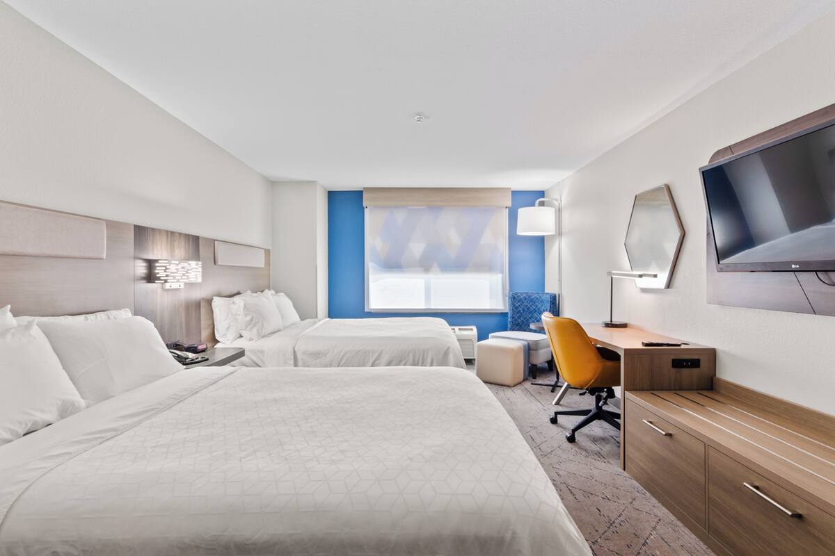 A hotel room with two beds, a desk, a chair, a TV, and a window with blinds.