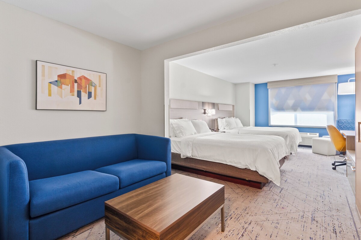 A modern hotel room with a blue sofa, a wooden coffee table, two beds with white bedding, a desk with a chair, and a large window with a view.
