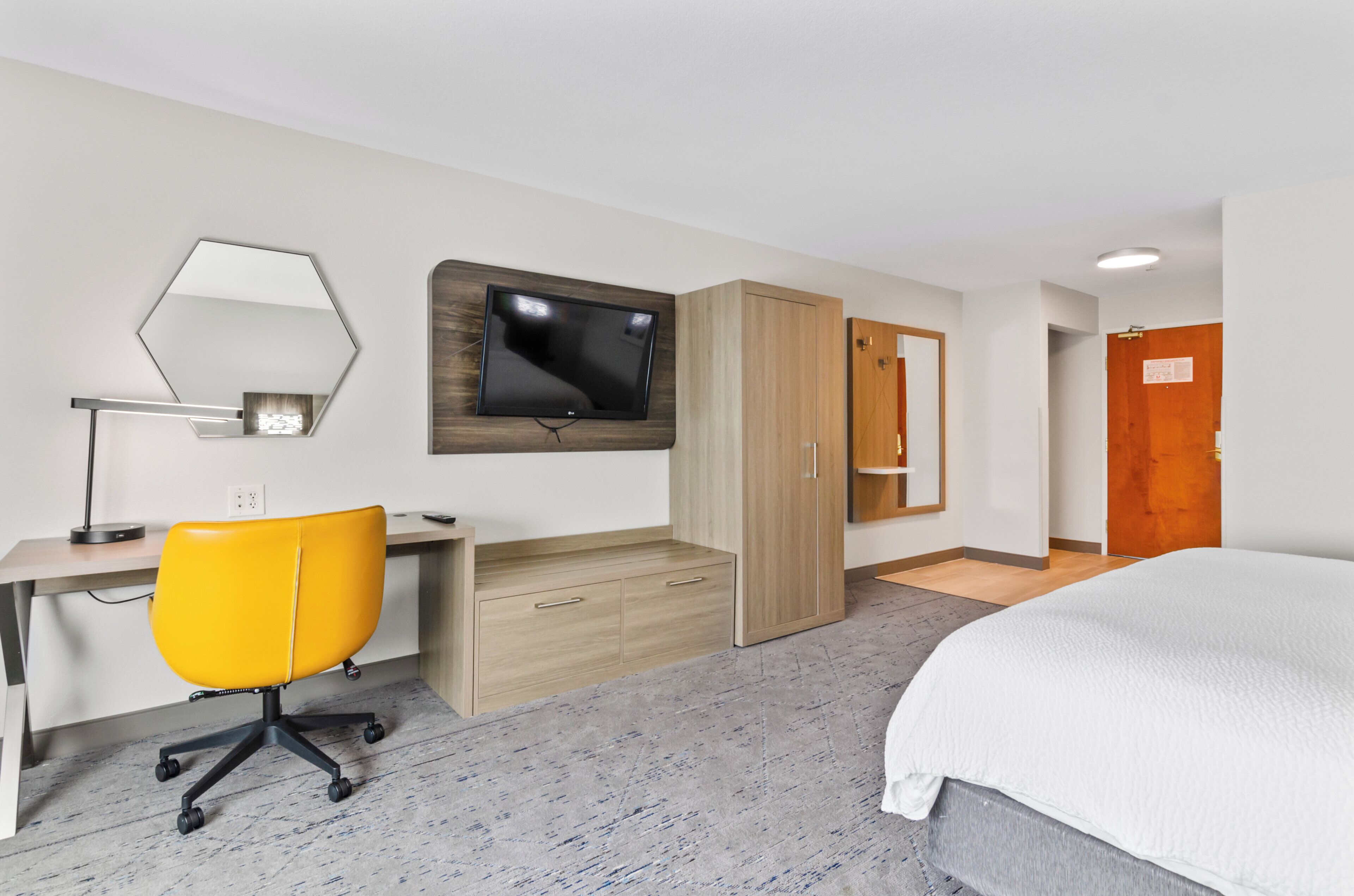 A hotel room with a bed, a desk with a chair, a TV mounted on the wall, and a wardrobe.