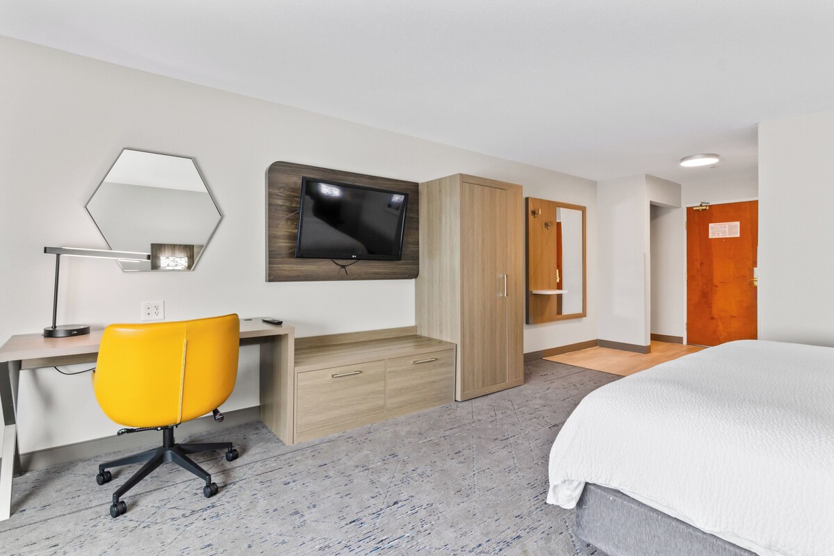 A hotel room with a bed, a desk with a chair, a TV mounted on the wall, and a wardrobe.