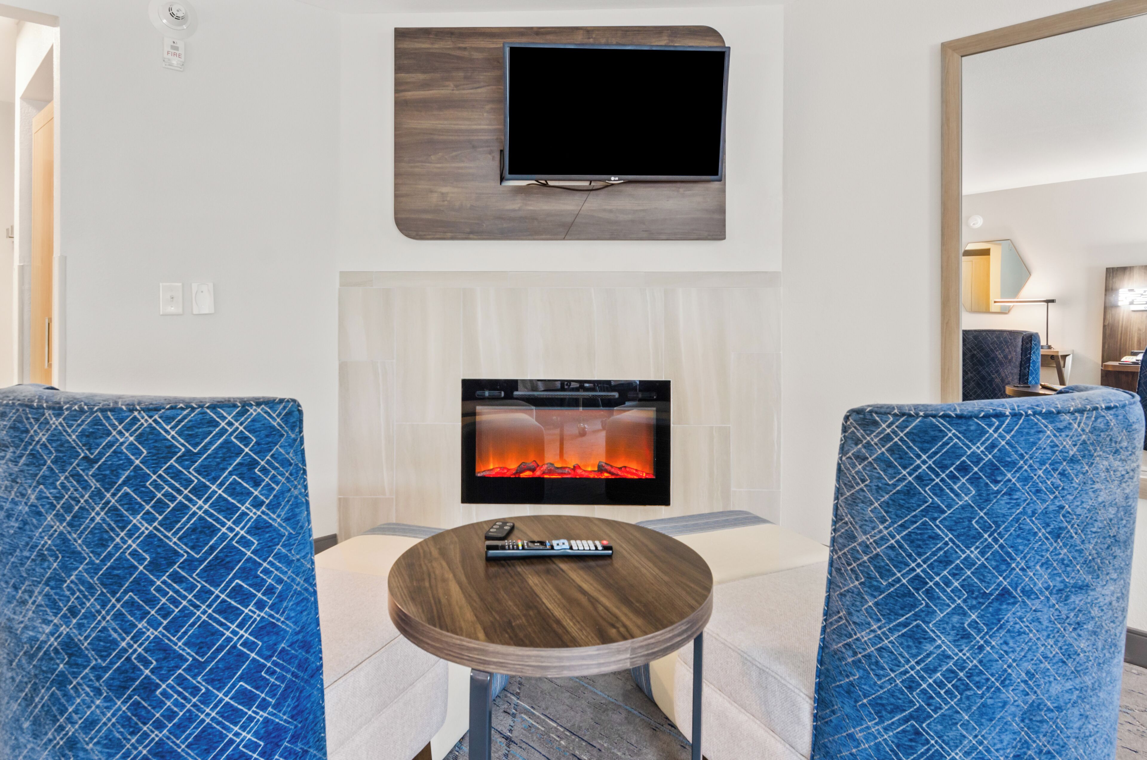Een moderne woonkamer met een flatscreen televisie aan de muur, een haard met een televisiescherm en twee blauwe fauteuils met een patroon.