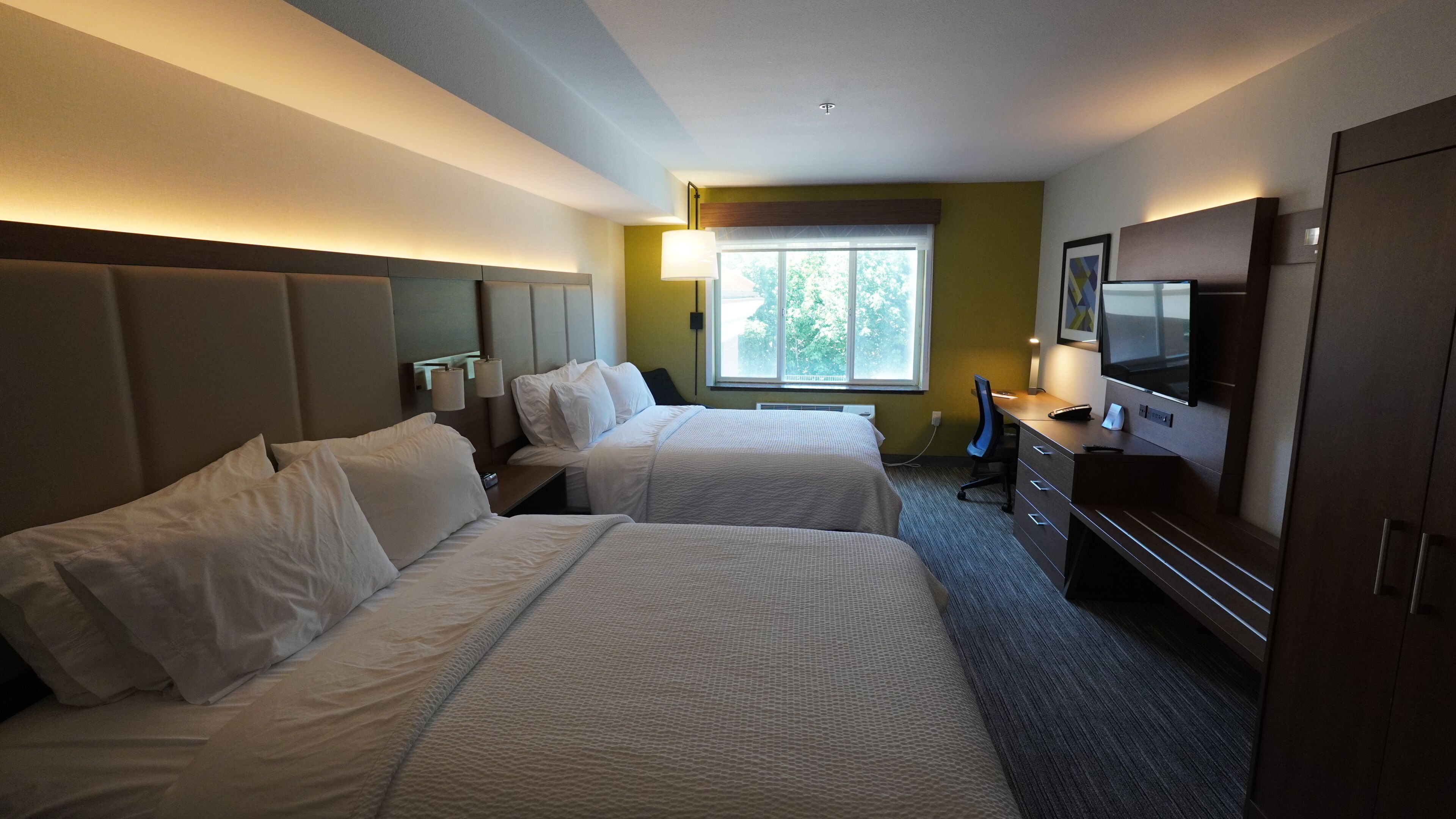 A hotel room with two beds, a desk with a computer, a TV, and a window with a view of trees.
