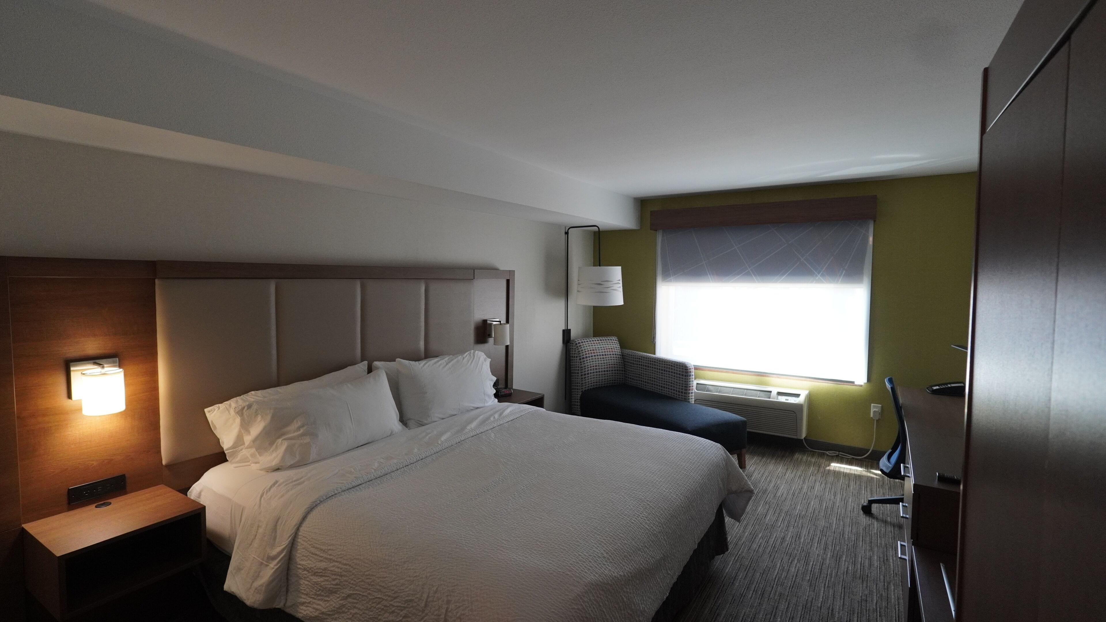 A hotel room with a large bed, bedside lamp, and a window with blinds.