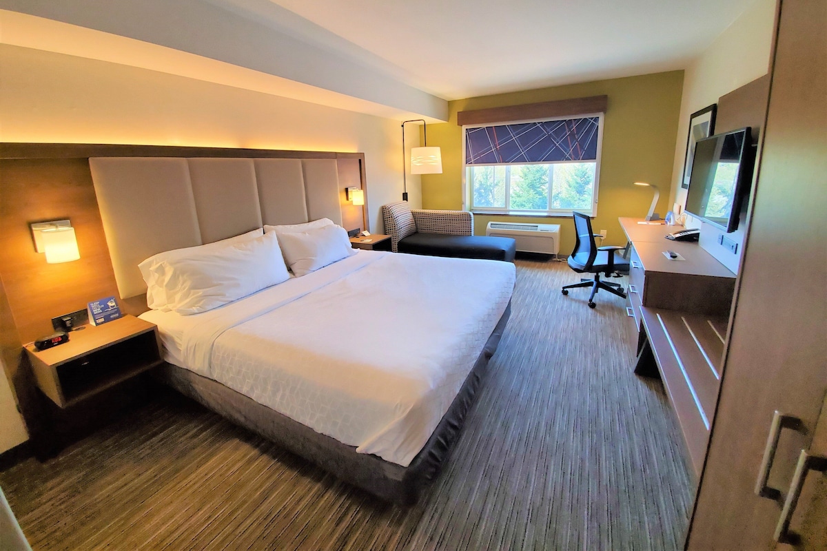 A hotel room with a large bed, a desk with a computer, a chair, a TV, and a window with a view.