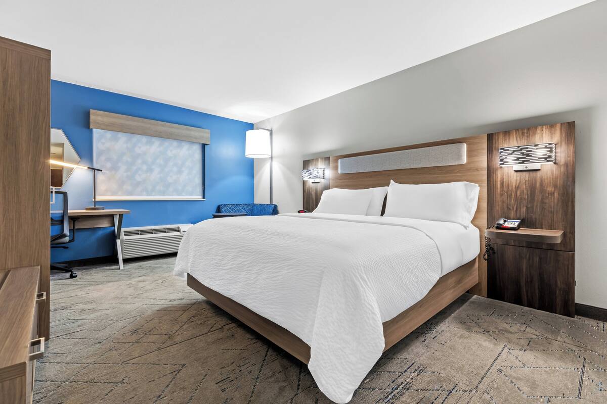 A hotel room with a large bed, a desk with a chair, a blue accent wall, and a modern headboard.