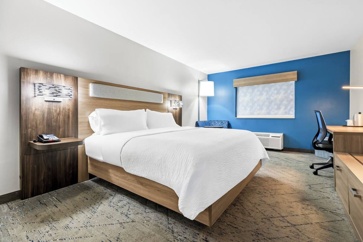 A hotel room with a bed, bedside table with a phone, a desk with a chair, and a window with blinds.