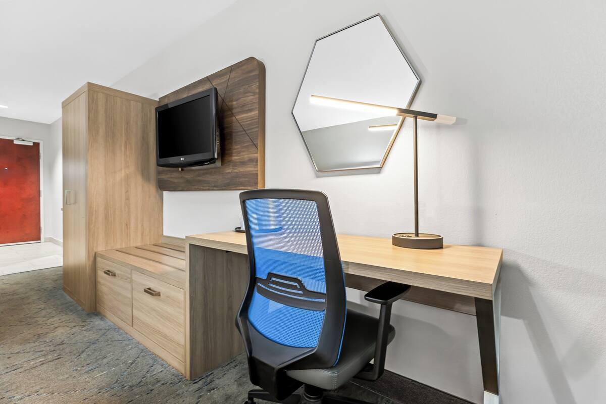 A modern office space with a wooden desk, a blue mesh office chair, a mounted flat-screen TV, and a wall-mounted desk lamp.
