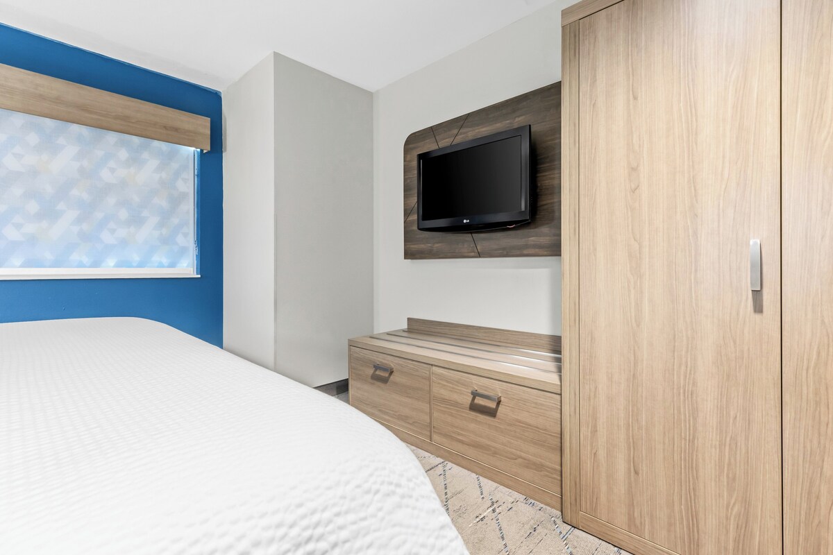 Une chambre à coucher comprenant un lit, un téléviseur fixé sur un panneau en bois, une armoire et une fenêtre avec des stores.