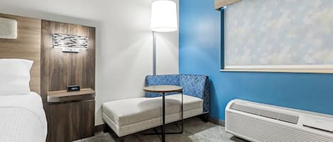 Quarto de hotel com cama, uma pequena mesa, cadeira, mesa de cabeceira, abajur e aquecedor fixado na parede.