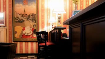 A hotel lobby with a checkered floor, striped wallpaper, a framed poster of a cityscape, a vase with flowers, and a lamp.