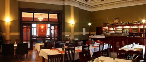 A restaurant with multiple tables and chairs, a bar area with shelves of bottles, and a large window with curtains.