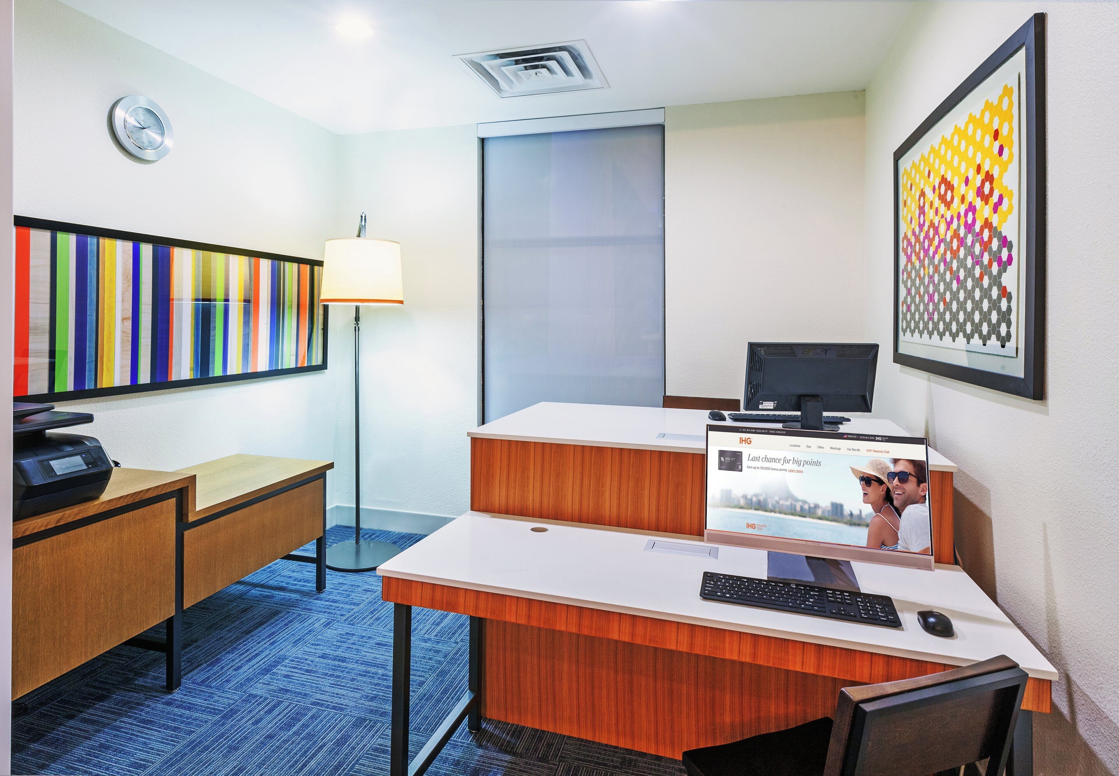 A hotel room with a desk, chair, computer, printer, lamp, and a large window.