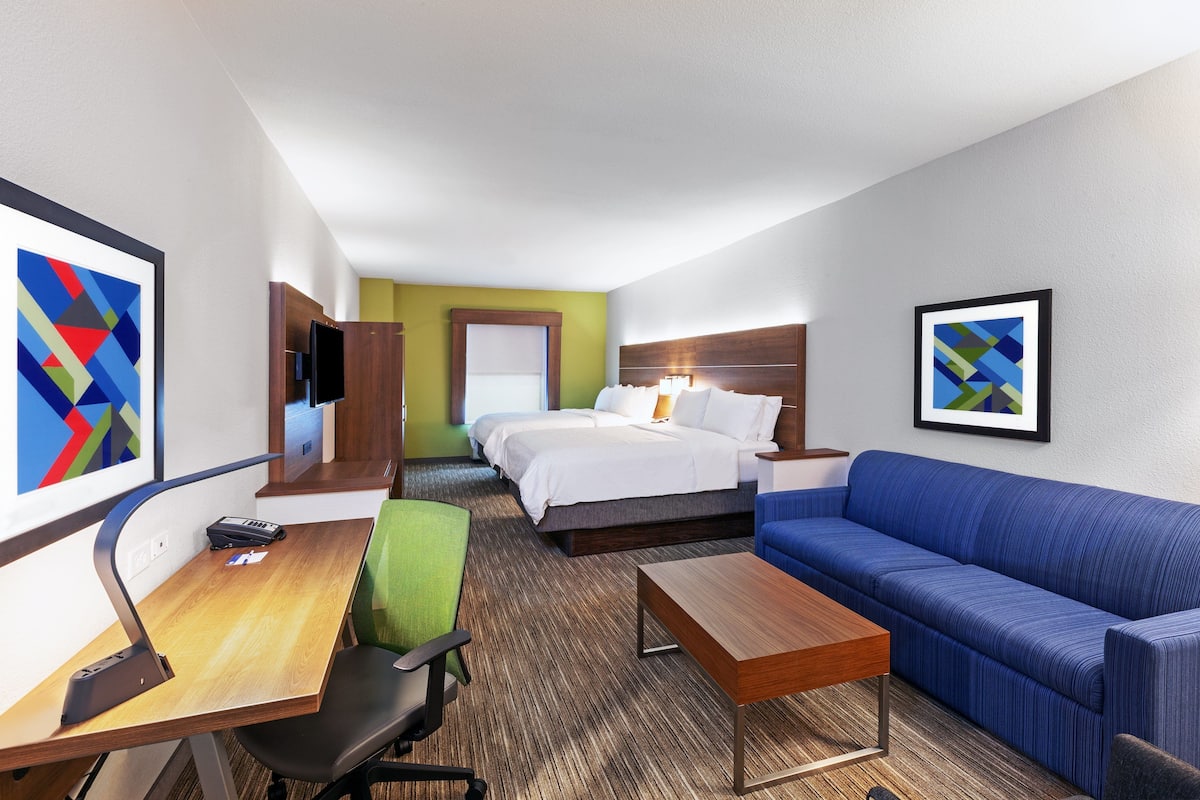 A hotel room with a bed, a blue sofa, a desk with a chair, a TV, and artwork on the walls.