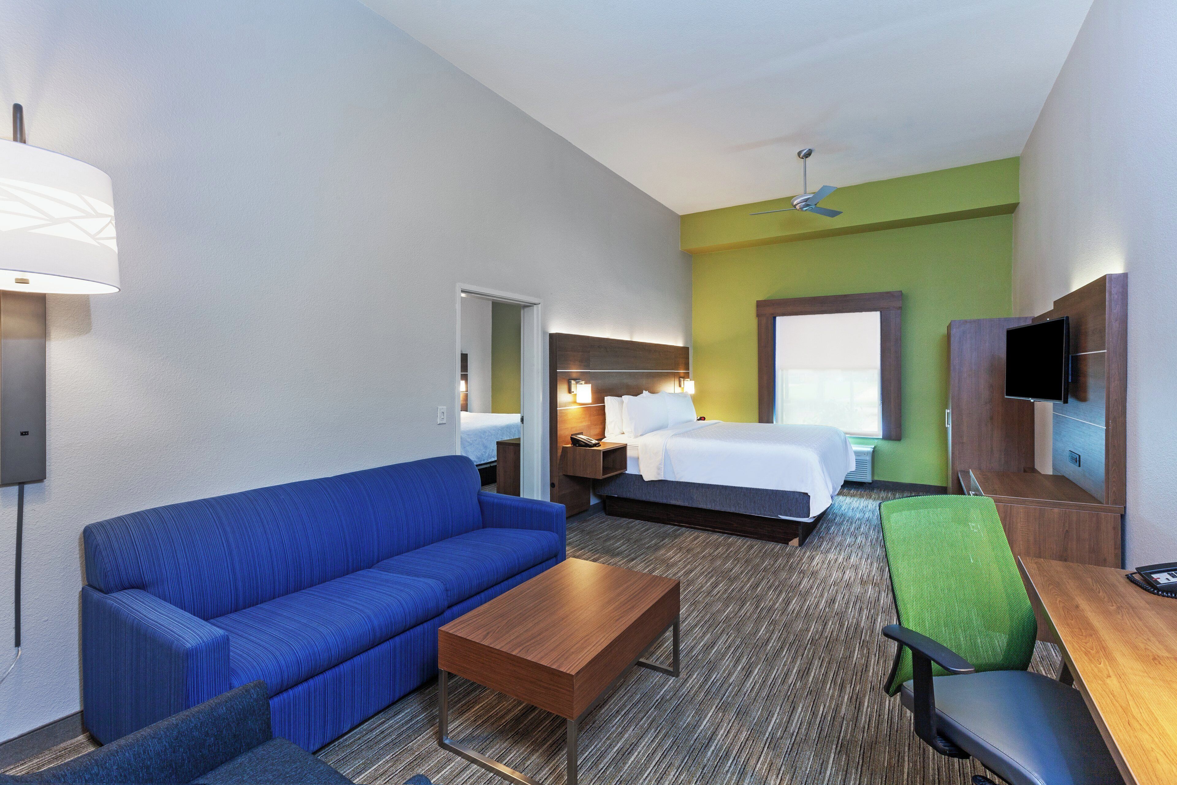 Habitación de hotel con sofá azul, una mesita de centro de madera, cama con ropa de cama blanca, una silla verde y un televisor de pantalla plana montado en la pared.