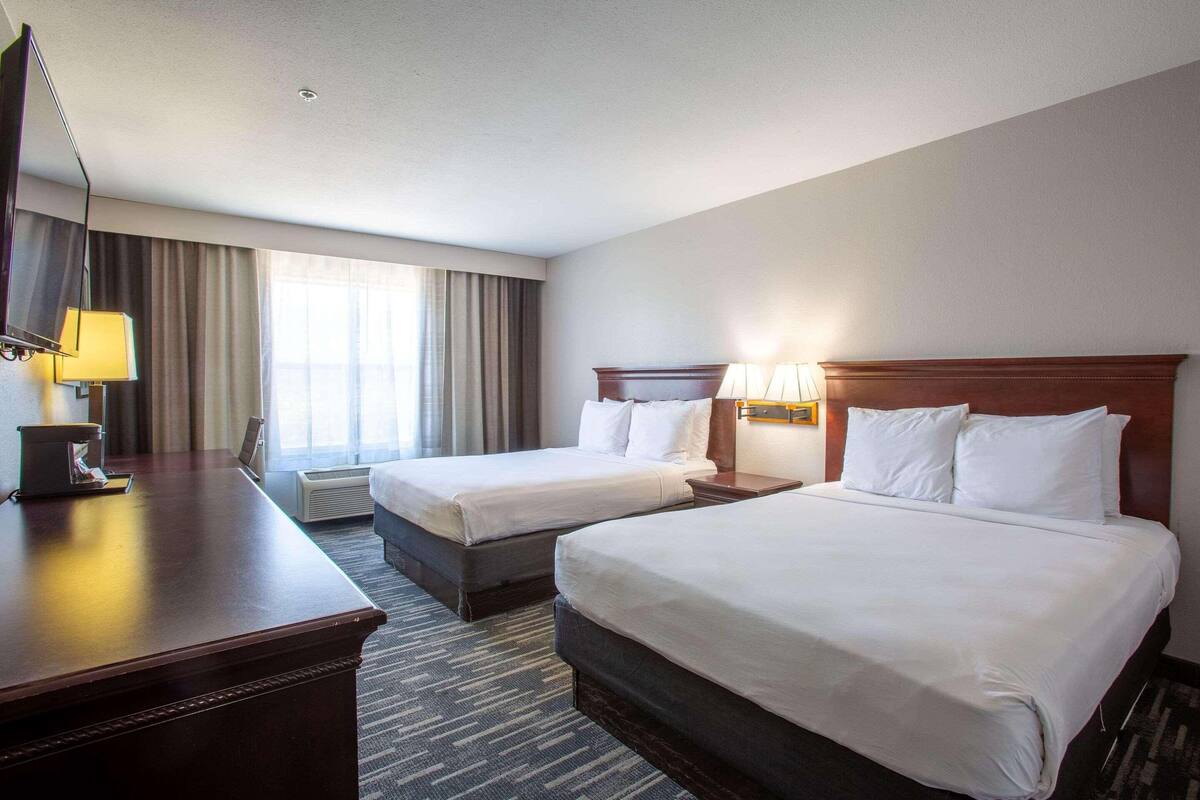 A hotel room with two beds, a desk, a lamp, and a window with curtains.