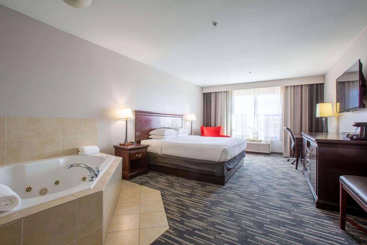 A hotel room with a large bathtub, a double bed, a desk with a chair, and a large window with curtains.