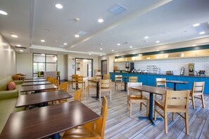 A modern dining area with multiple tables and chairs, a kitchen area with blue cabinets, and a seating area with a green couch. - Country Inn & Suites by Radisson, Tucson City Center, AZ (Tucson)
